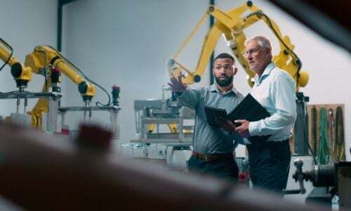 Two men review operations on a manufacturing floor with industrial robotic arms, representing veteran entrepreneur training and business growth in Florida's manufacturing sector. Two men review operations on a manufacturing floor with industrial robotic arms, representing veteran entrepreneur training and business growth in Florida's manufacturing sector.