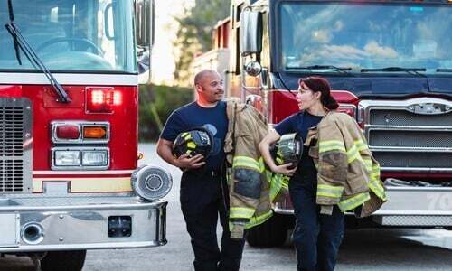 A male and female veteran in firefighter gear stand together outside a fire truck, representing veteran career placement with Florida public safety employers.