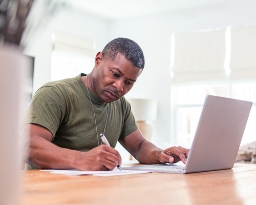 A veteran sits at a desk writing notes beside a laptop, representing Veterans Florida's Getting Started Cohort teaching new veteran-owned business registration, marketing, and accounting in Florida.